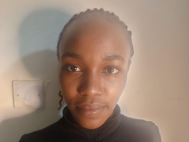 CocoaEclipse with brown skin, small nose ring, and braided hair, wearing a black zip-up, has a subtle smile, and is looking at the camera with soft, brown eyes.