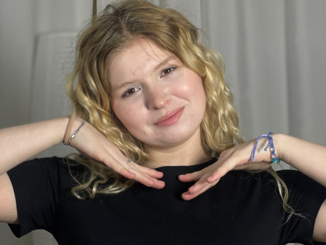 Delphine Hambleton hat blondes, lockiges Haar, helle Haut und ein leichtes Lächeln. Sie trägt ein schwarzes Hemd, ihre Hände liegen in der Nähe ihres Halses, und an ihrem rechten Handgelenk trägt sie blaue Armbänder..