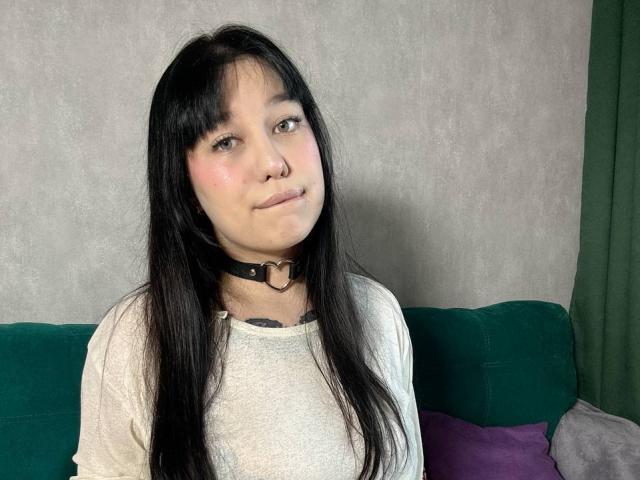 AnieEva with long black hair, pink cheeks, and a small nose ring, wearing a black choker with a heart ring, and a white shirt, sitting on a green couch.
