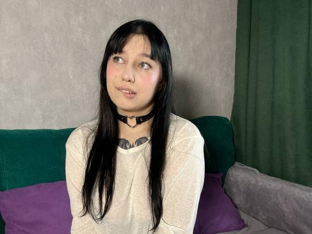 AnieEva with long black hair, wearing a black choker and beige shirt, sitting on a green and purple sofa, looking up with a slight smile, showing her tits.