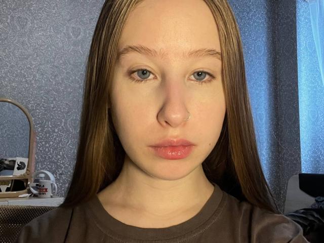 CorieCecchetti has pale skin, blue eyes, and a nose ring. She's pouting with glossy pink lips, wearing a brown shirt, and has long, straight brown hair.