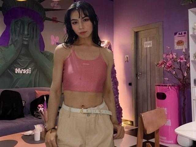 KatherinJohns with dark wet hair, pink sports bra showing small tits, beige high-waist pants, white belt, standing in purple-lit room, pink vase, green Buddha painting in background.