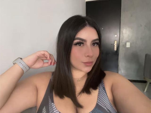 GabrielaFerrer with medium brown bob, wearing a gray sports top, showing her large boobs, red nails, and a smartwatch, standing near a black door.