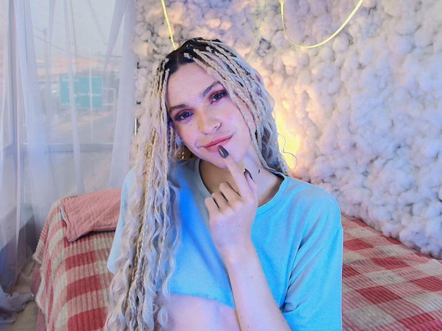 AaronFinn with long blonde braids, pink eye makeup, and a blue crop top, touching his chin with a finger, smiling, on a red checkered bed.