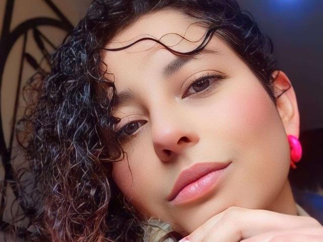 IrinaSweett's face, with curly black hair, pink earrings, and soft pink lipstick, looking slightly to the side with a subtle smile. Her skin is smooth and light brown.