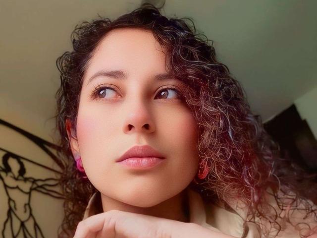 IrinaSweett has curly brown hair, pink earrings, and pink lips, gazing up with brown eyes, her hand under her chin, wearing a beige shirt, in a soft light.