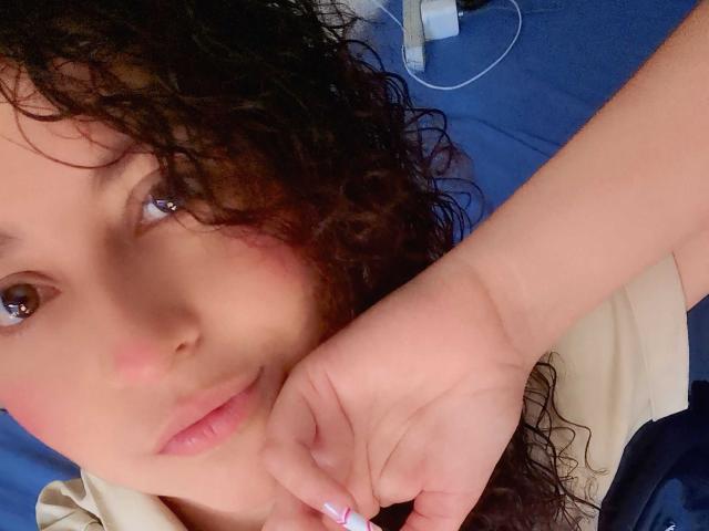 IrinaSweett's face with curly black hair, brown eyes, and pink lips, resting her chin on her hand with pink nails, wearing a beige shirt, on blue bedding.