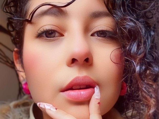 IrinaSweett's face, curly dark hair, pink lips, brown eyes, pink earrings, finger on lower lip, glossy pink nails, soft skin, slightly parted lips, sensual expression.