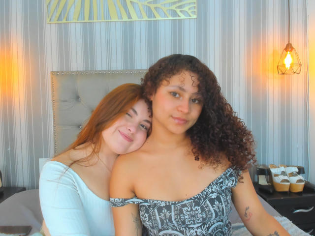 SofyViky with red hair and white top, leans on SofyViky with curly brown hair and black floral dress, showing her tits, in a cozy bedroom with warm lights.