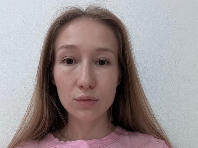 AlyssaKampe has long brown hair, light skin, small nose, and neutral expression. She's wearing a pink shirt, with her boobs partially visible, against a white textured wall.