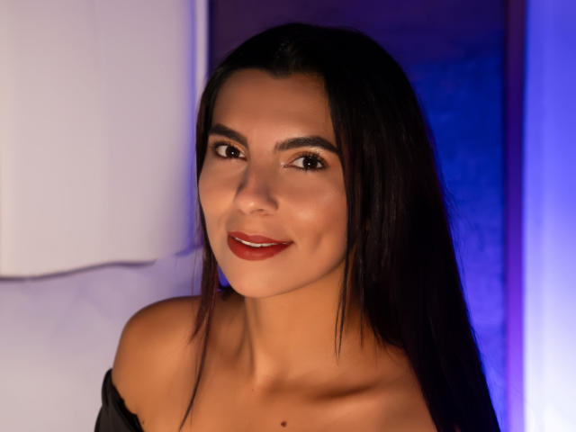 CarlaTorres has long dark hair, red lipstick, and a cute smile. She's wearing an off-shoulder black top, showing her collarbone and a small mole. She's looking at the camera.