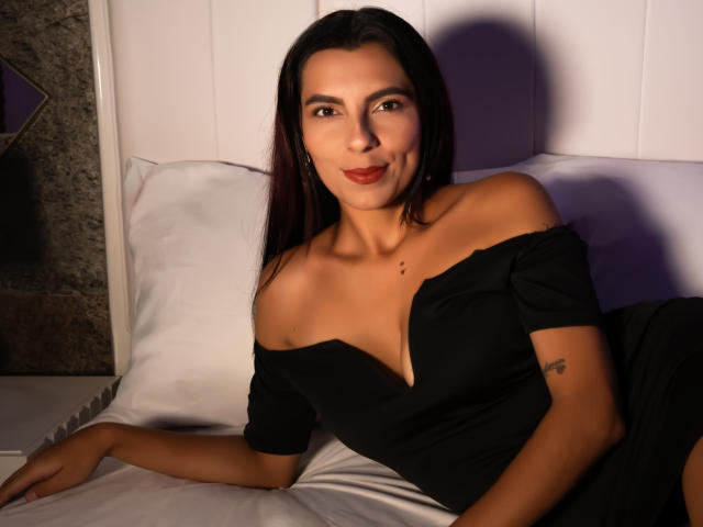 Carla Torres, com cabelo escuro, vestindo um vestido preto com um ombro descoberto, sentada em uma cama, sorrindo, mostrando seios pequenos, uma tatuagem no braço e uma pequena pinta no peito..