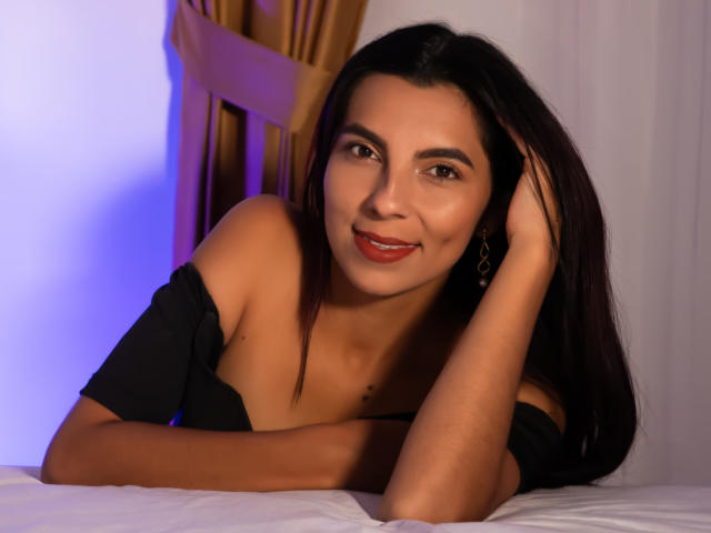CarlaTorres tiene el pelo oscuro, ojos marrones, lápiz labial rojo y una sonrisa encantadora. Está acostada en una cama con una blusa negra con un hombro descubierto, con una mano en la cabeza, mirando a la cámara..