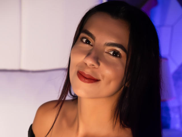 Carla Torres, con cabello largo y oscuro, ojos marrones y lápiz labial rojo, sonríe suavemente a la cámara. Lleva una blusa con un hombro descubierto, que muestra un poco de su clavícula..