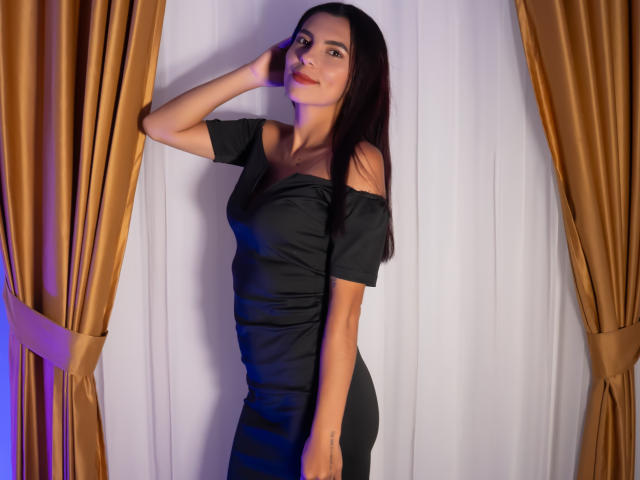 CarlaTorres with long dark hair, black off-shoulder dress, standing, hand on head, looking at camera, smiling, brown curtains on both sides, white curtain background.