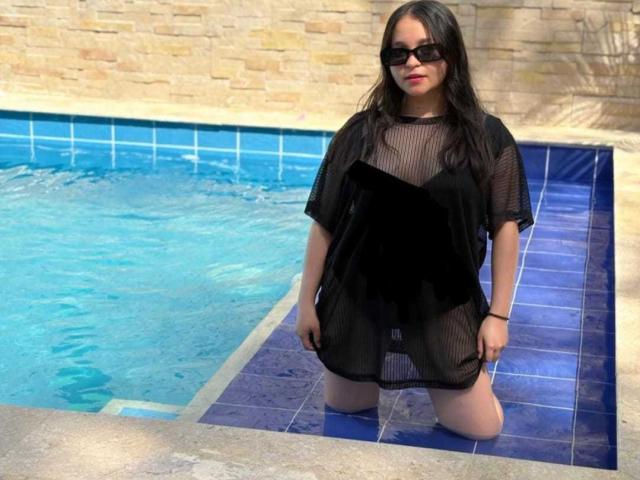 SamyMancini kneels in a blue-tiled pool, wearing a black mesh dress that reveals her pussy and black panties, with black sunglasses and long black hair.