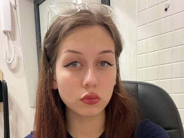 A RussellMixer tem olhos verde-claros, delineador escuro e batom rosa. Ela está usando óculos transparentes na cabeça, tem cabelo castanho e uma expressão séria. Seios não visíveis..