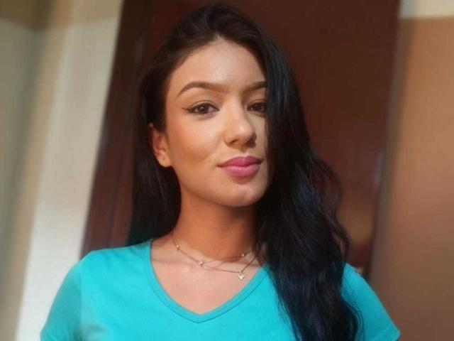 IvorySelene has dark, wavy hair, tan skin, pink lipstick, and is wearing a teal shirt with a delicate necklace. Her face is slightly turned, showing a subtle smile.