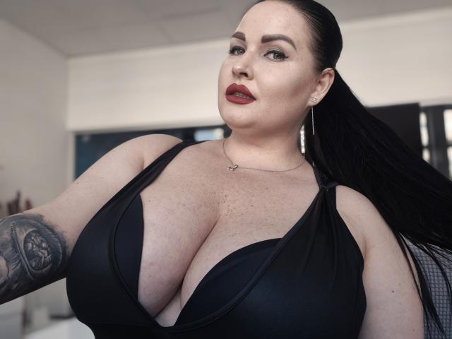 GeorgiaLerox with large, round boobs in a black, deep V-neck top, fair skin, red lipstick, black tattoo on arm, long black hair, and a confident, sexy look.