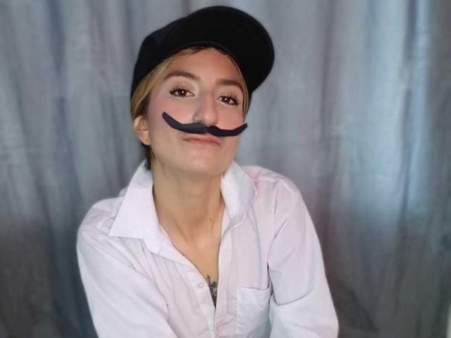 Kaiix with a black mustache sticker, wearing a black cap and white shirt, smiling slightly. Short blonde hair, light skin, subtle makeup. Boob outline visible under shirt.