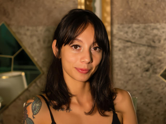 Sara Lambert tiene una piel marrón media, cabello negro de largo medio, pechos pequeños, un tatuaje en el hombro derecho y una ligera sonrisa, y lleva puesto un top negro con tirantes finos..