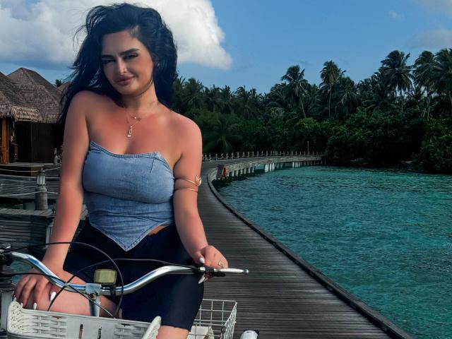 NightNymph with black hair, blue strapless top, and black shorts, sitting on a bike on a wooden dock, ocean and palm trees in the background, smiling, hands on bike handles.