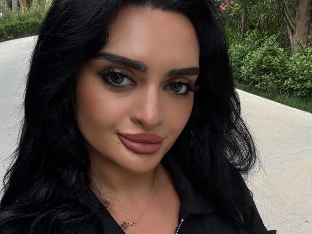 NightNymph hat langes, schwarzes Haar, dunkle Augen und rosafarbene Lippen. Sie trägt ein schwarzes Oberteil und blickt mit einem leichten Lächeln in die Kamera. Ihre Brüste sind nicht sichtbar..