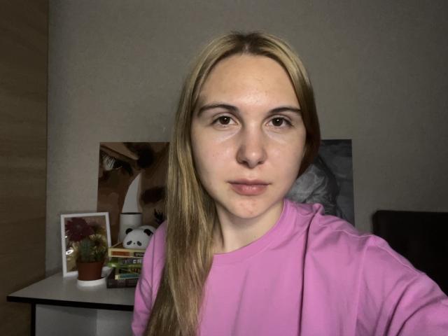 DeeannMilloy has blonde hair, pink shirt, and a neutral expression. She's in a dim room with a potted plant and small panda figurine in the background.