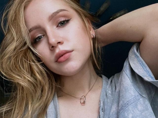 JanellLindinha has blonde hair, light skin, and is wearing a striped shirt with a necklace. She's looking at the camera with her hand behind her head.