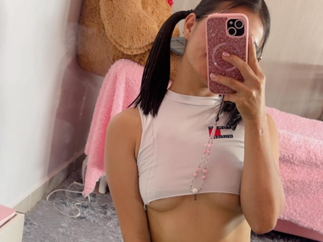 KimmyK with pigtails, wearing a white cropped top showing her tits, holding a pink glitter phone, pink beads around her neck, taking a mirror selfie.