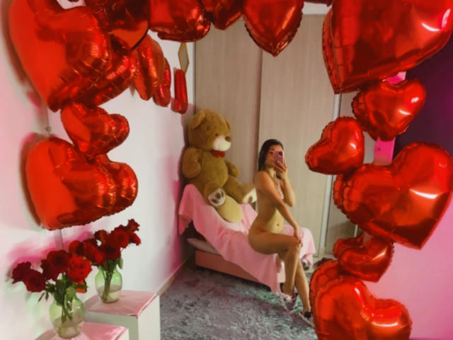 KimmyK, nude, sitting on pink bed, next to giant brown teddy bear, surrounded by red heart balloons, holding phone, with red roses on table, in pink room.