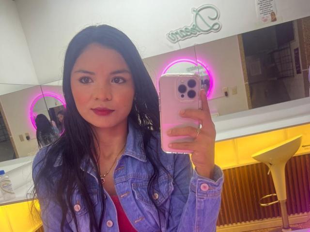 CrystalStones with long black hair, red lipstick, and a denim jacket, holding a pink phone, taking a mirror selfie, pink neon lights in the background.