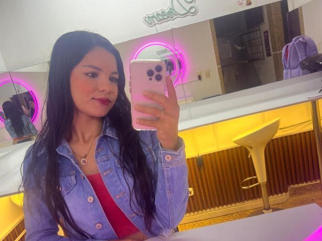 CrystalStones with long black hair, wearing a blue denim jacket and red top, taking a mirror selfie with pink neon rings, holding a pink iPhone, smiling slightly.