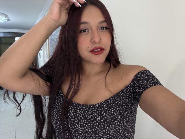 MarianaVargasX with long dark brown hair, red lipstick, nose ring, wearing a black floral off-shoulder top, hand in hair, slightly smiling, light skin, medium-sized tits.