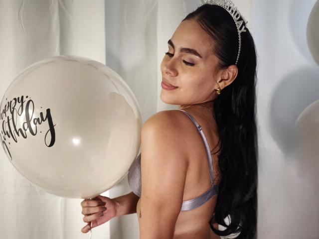 RossaHot with long black hair in a silver bra, holding a white birthday balloon, wearing a tiara, smiling, eyes closed, showing her taut ass and small gold earrings.