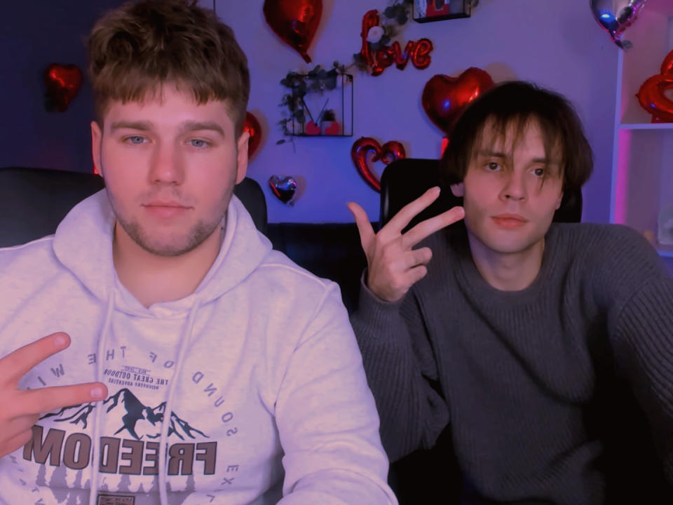 MichaelAlexCpl with a white hoodie and another MichaelAlexCpl with a gray sweater, both making peace signs, in a room with red heart balloons and blue lighting.