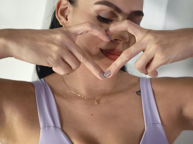 RossaHot with pale skin and dark hair, making a heart shape with her fingers, wearing a lilac top, gold necklace, and a small tattoo on her right shoulder.