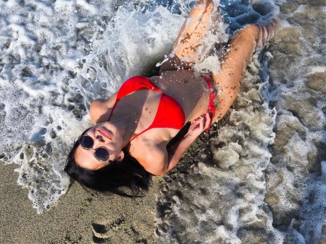 RossaHot with red bikini, wet skin, and black hair, lying on beach, waves washing over her, wearing round sunglasses, her tits and ass partially visible, water splashing.