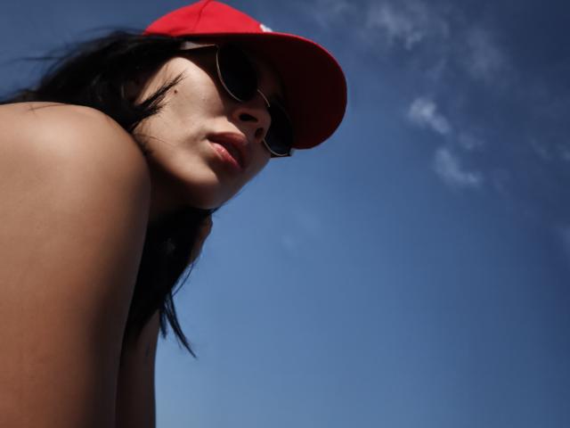 RossaHot with dark hair, red cap, and sunglasses, looking down with pouty lips, showing bare shoulders, against a blue sky. Her tits are not visible.
