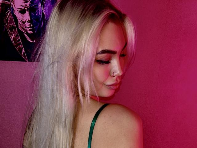 LexiLee with blonde hair, green strap, nose ring, pink and purple lights, looking down, showing her ass, eyelashes, and a subtle smile. Her tits are partially visible.