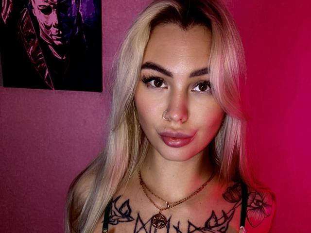 LexiLee with blonde hair, nose ring, and tattoos on her tits, wearing a necklace, against a pink background. Her eyes are brown, and she's giving a slight smirk.