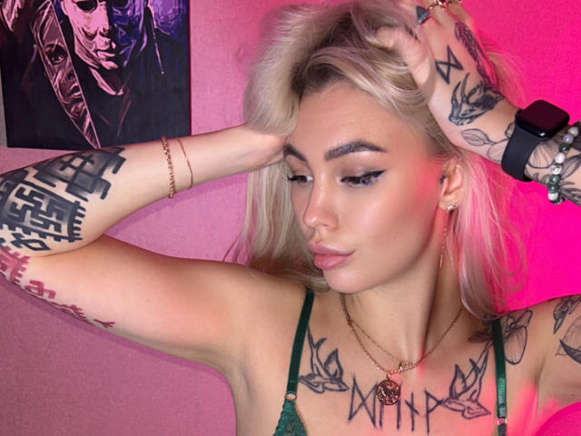 LexiLee with blonde hair, green bra, tattoos on arms and chest, wearing a black smartwatch, pink lighting, hand in hair, subtle makeup, pink wall, confident expression.