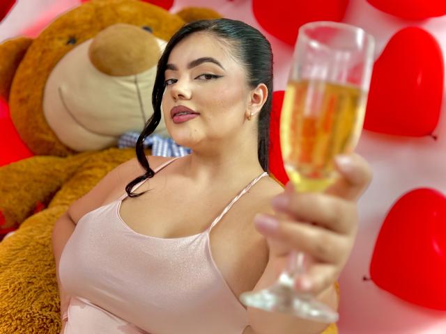 LaraJames with big tits in a pink tank top, holding a champagne glass, tongue out, black hair in a ponytail, sitting next to a giant teddy bear.
