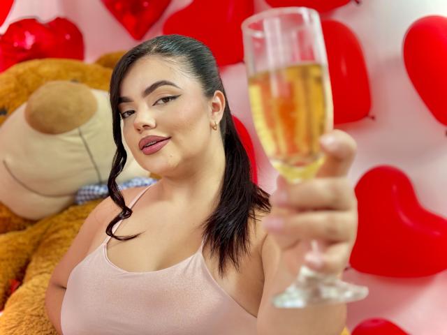 LaraJames with dark hair in a ponytail, pink tank top, holding a glass of champagne, smiling, showing her tits, red heart balloons, and a big brown teddy bear in background.