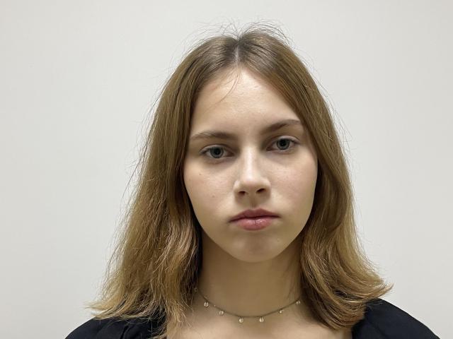 IoneTrosper has shoulder-length brown hair, fair skin, blue eyes, and a neutral expression. She wears a black top and a delicate necklace with small beads.