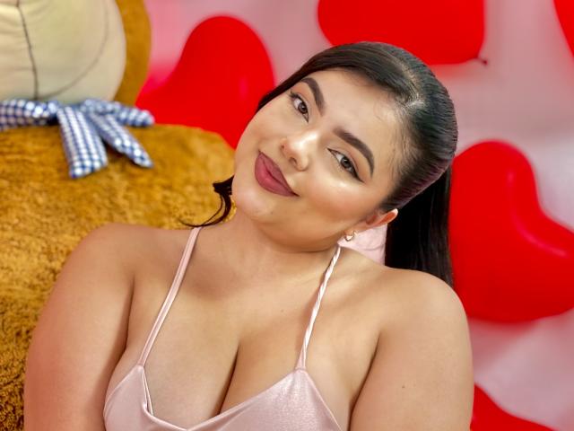 LaraJames with dark hair in a ponytail, wearing a pink satin top, showing her tits, smiling at the camera, pink lipstick, brown eyes, red heart-shaped background.