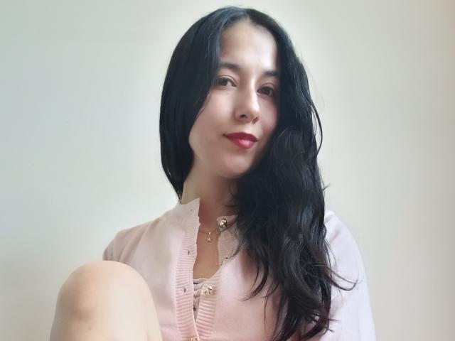 AnneStone with long black hair, pink button-up shirt, and red lipstick, sitting with one knee up, smiling softly, wearing a gold necklace with small pendants.