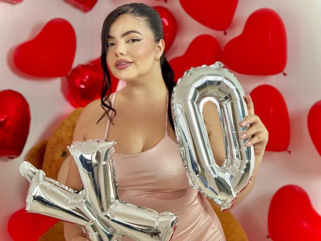 LaraJames with dark hair in a ponytail, wearing a pink satin dress, holding shiny silver "10" balloons, red heart balloons in the background, smiling with pink lipstick.