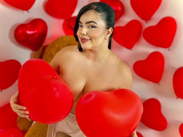 LaraJames with big boobs, holding red heart balloons, smiling, wearing a strapless white dress, dark hair in a side braid, pink lipstick, gold hoop earrings, warm light.