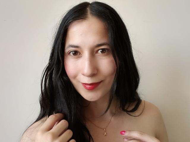 AnneStone has long black hair, brown eyes, and red lipstick. She's smiling with pink nails, holding her hair, wearing a gold necklace, and has a cute, sexy face.